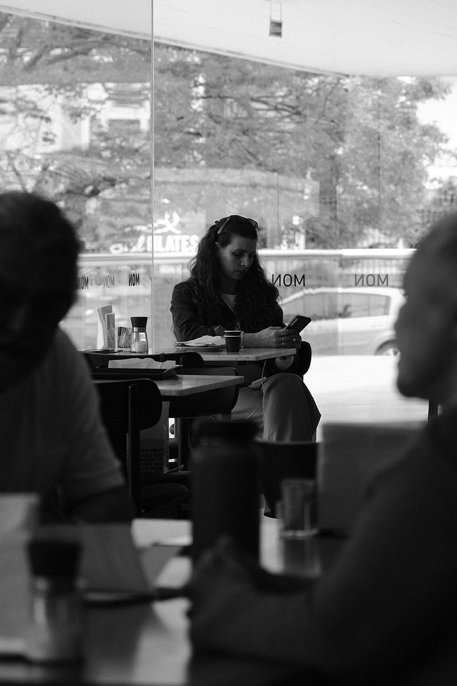 A woman sitting at a café table looking at her phone, seen through a window with silhouetted figures in the foreground.